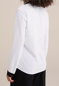 White long-sleeve shirt with a black collar and buttoned cuffs. Features a textured fabric with a relaxed, slightly curved hem.