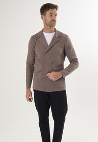 Bruine gebreide cardigan met een diagonale textuur, dubbelgeknoopte knopen en een gelapte kraag. Gecombineerd met zwarte broek.
