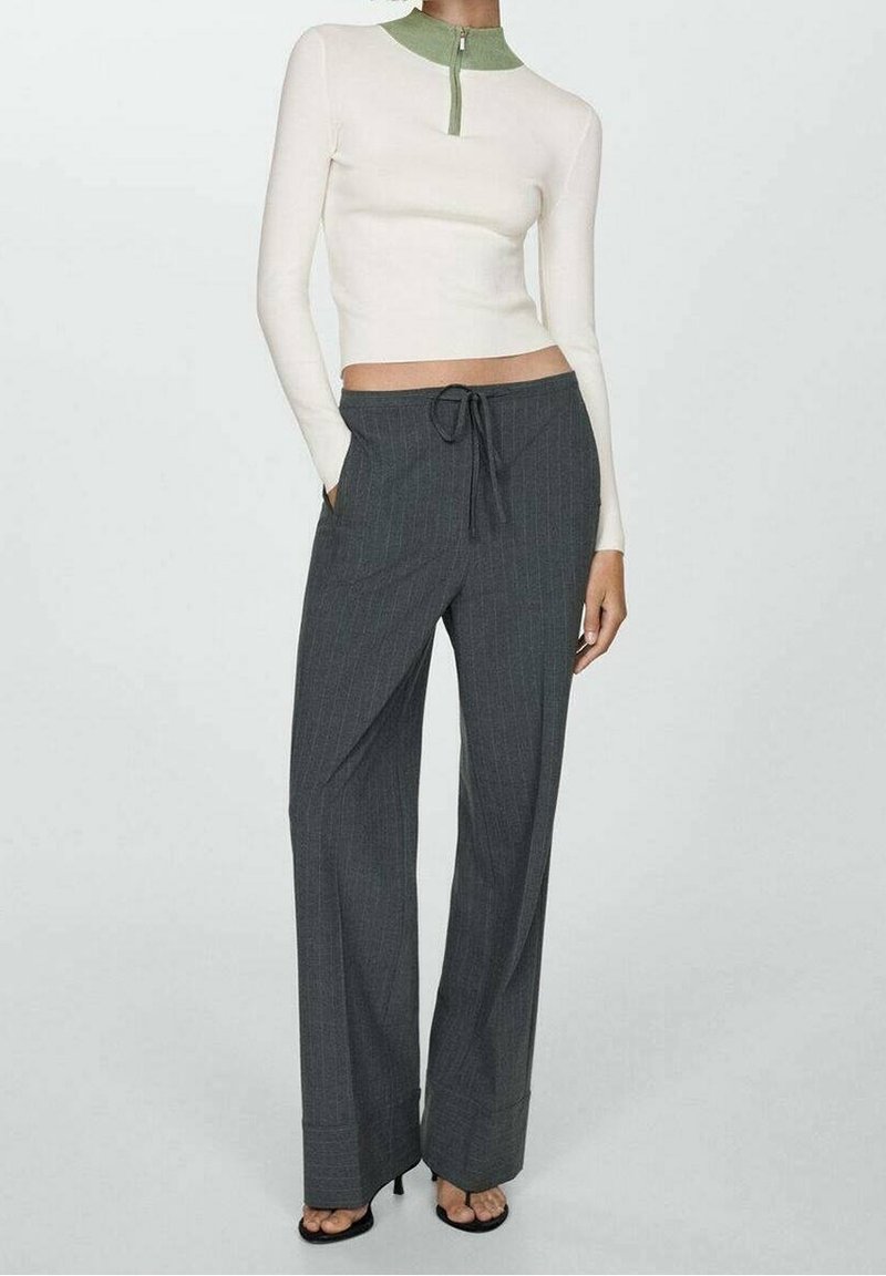 Haut à manches longues crème avec col vert et fermeture éclair, associé à un pantalon large à rayures fines gris foncé avec une ceinture ajustable.