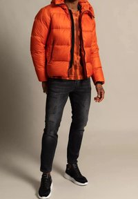 Orange puffjacka med huva, dragkedja framtill och sidofickor, i kombination med en mönstrad skjorta och svarta jeans. Svarta sneakers.