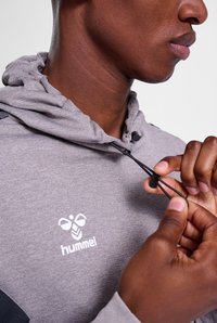 Felpa grigia con zip, realizzata in un tessuto textured, con accenti neri sulle spalle e un logo sul petto; dotata di un cappuccio regolabile con lacci.