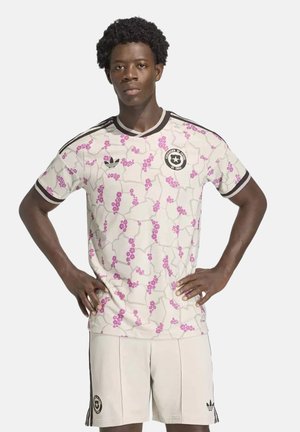 Jeune homme aux cheveux courts et bouclés porte un maillot de football blanc et un short avec un motif floral rose et une bordure noire, debout les mains sur les hanches.