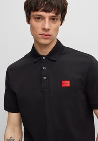 Black polo shirt with a ribbed collar, three buttons, and a red rectangular logo patch labeled 'HUGO' on the left chest.