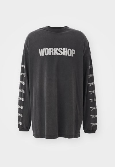 Svart langermet T-skjorte laget av myk bomull, med ordet "WORKSHOP" trykt i sølv på forsiden og gjentatte grafikker på ermene.