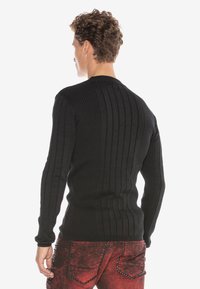Schwarzer gerippter Strickpullover mit langen Ärmeln, enganliegendem Design, runder Halslinie und strukturierter Oberfläche, kombiniert mit roten Jeans mit Nieten.