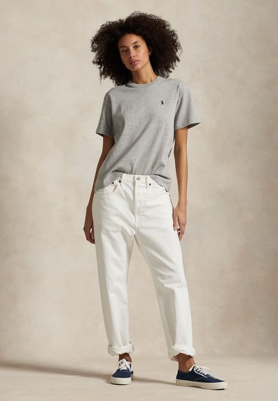 Žena s kučeravými vlasmi, ktorá má na sebe sivé tričko s krátkym rukávom, biele vyhrnuté nohavice a tmavomodré tenisky, stojí pred béžovým pozadím.