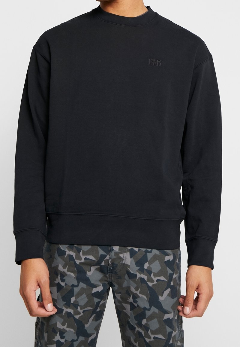 Uomo che indossa un semplice felpa nera Levi's e pantaloni mimetici grigio-blu, in piedi con le braccia rilassate ai lati.