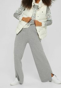 Eine Frau trägt graue weite Hosen mit seitlichen Schlitzen, weiße Sneakers, einen gemusterten grauen Pullover und eine weiße Daunenweste und stellt den Reißverschluss der Weste ein.