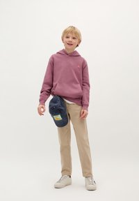 Jongen in een mauve hoodie met een klein logo, beige broek en witte sneakers, die een marineblauwe pet met een kleurrijke patch vasthoudt.