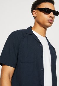 Camisa de manga corta, de color azul marino oscuro, con una tela texturizada, un bolsillo en el pecho y botones negros, superpuesta sobre una camiseta blanca de cuello redondo.