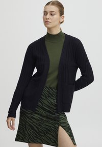 Mörkblå ribbad cardigan med två framfickor, bärs över en grön polokrage och en grön kjol med tigerränder och knappar.