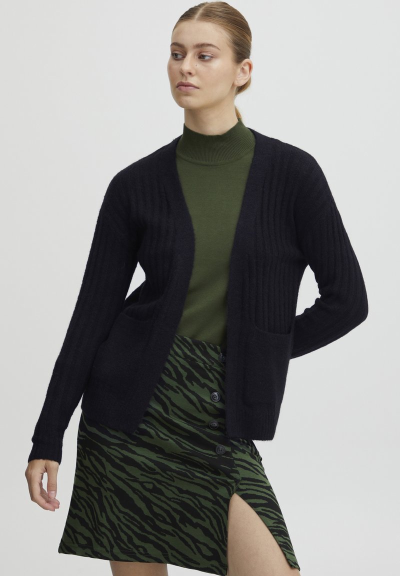 Mörkblå ribbad cardigan med två framfickor, bärs över en grön polokrage och en grön kjol med tigerränder och knappar.