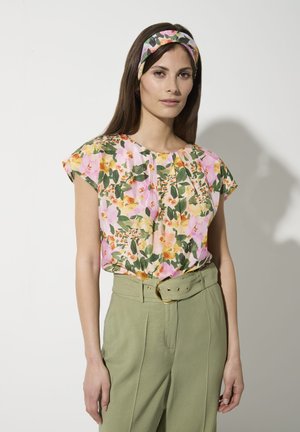 Blumenbluse in Pink mit gelben und grünen Mustern, kurze Ärmel, plissierter Vorderteil. Kombiniert mit hochtaillierten, hellgrünen Hosen und einem passenden Haarband.