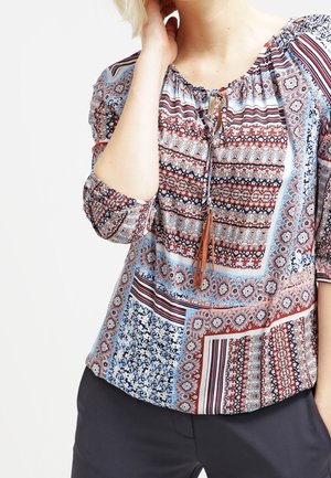 Vrouw draagt een meerkleurige blouse met patroon, franjekoord bij de halslijn en driekwart mouwen, gecombineerd met donkere broek.