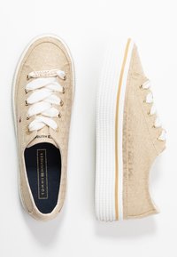 Sneakers in tela beige con lacci bianchi e suole in gomma. Accenti dorati sui fori dei lacci e un patch con logo sulla linguetta. Superficie texture.