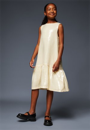 Cream sequined dress with a flared hem and sleeveless design. Paired with black shoes featuring a strap and thick sole.