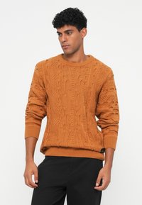 Πλεκτό πουλόβερ σε καμένο πορτοκαλί με υφασμάτινα καλωδιακά σχέδια, ριγέ μανσέτες και στρογγυλή λαιμόκοψη, συνδυασμένο με μαύρο παντελόνι.