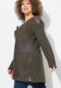 Cardigan verde oliva a maglia con vestibilità ampia, caratterizzato da una texture a coste, chiusura con lacci e spacchi laterali. Indossato sopra un top rosa a fantasia.