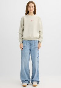 Sudadera gris claro con logo rojo bordado, combinada con vaqueros de pierna ancha azul claro con bolsillos laterales estilo cargo. Unas zapatillas blancas completan el conjunto.