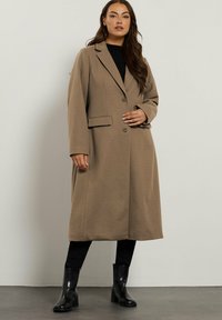 Donna che indossa un lungo cappotto beige sopra pantaloni neri e stivali con tacco neri, in piedi contro una parete bianca semplice.