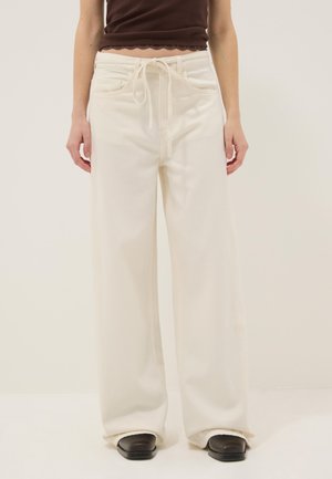 Donna che indossa pantaloni bianchi a gamba larga con vita alta e coulisse, top marrone corto e scarpe nere con punta quadrata.