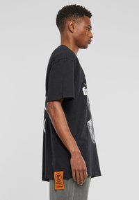 Melna kokvilnas t-kreklā ar īsām piedurknēm, ar grafisku apdruku priekšpusē un izteiksmīgu oranžu birku sānos.