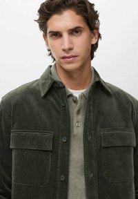 Joven con cabello castaño ondulado, vistiendo una chaqueta de pana verde oscura sobre una camisa gris abotonada, mirando a la cámara con una expresión neutral.