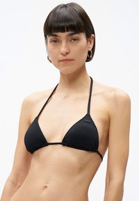 Mladá žena s krátkymi hnedými vlasmi, ktorá nosí zlaté kruhové náušnice a čierny textúrovaný trojuholníkový bikínový vrch na jednoduchom bielom pozadí.