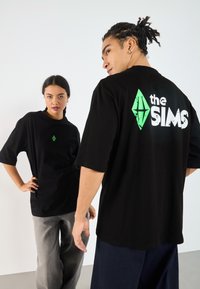 Svarte unisex t-skjorter med en grønn Sims-logo på forsiden og hvit tekst på baksiden. Løs passform, korte ermer, bomullsmateriale.