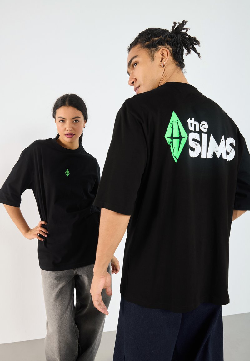 Svarte unisex t-skjorter med en grønn Sims-logo på forsiden og hvit tekst på baksiden. Løs passform, korte ermer, bomullsmateriale.