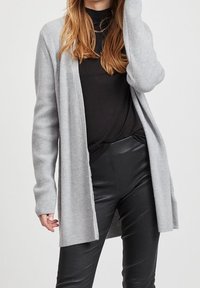 Femme portant un long cardigan tricoté gris clair sur un col roulé noir et un pantalon en cuir noir, posant avec une main près de sa tête.