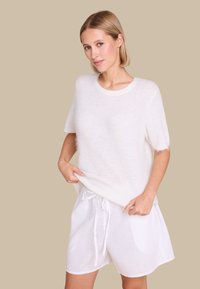 Lett, hvit, fuzzy strikkegenser med korte ermer, sammen med løse hvite shorts med snøring mot en nøytral beige bakgrunn.