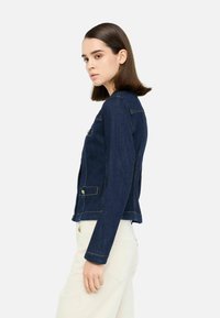 Džínová bunda, tmavě modrá, přiléhavý design se zlatými knoflíky, dvěma předními kapsami a jemným šitím, viditelná z boku.