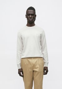 Muž stojaci pred bielym pozadím, oble�čený v krémovom pulóvri s okrúhlym výstrihom a béžových plisovaných nohaviciach, smerujúci dopredu s neutrálnym výrazom.