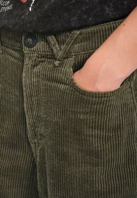 Pantaloni in velluto a coste verde oliva con righe verticali, dotati di una tasca frontale e chiusura a bottone nero, che mostrano una texture morbida.