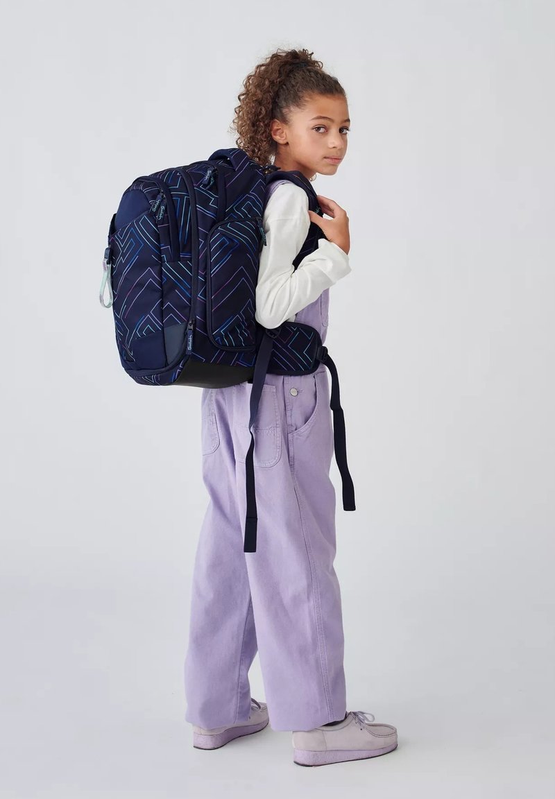 Mochila marina con un patrón geométrico en azul y púrpura, correas acolchadas, bolsillos laterales y asa superior; llevada por un niño con un overol lavanda.