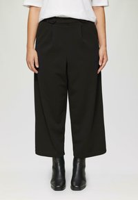 Zwarte wijde broek met twee voorplooien en zijzakken, gemaakt van een gladde stof. Draagt een paar zwarte enkellaarsjes.