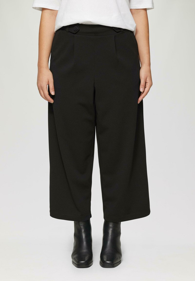 Zwarte wijde broek met twee voorplooien en zijzakken, gemaakt van een gladde stof. Draagt een paar zwarte enkellaarsjes.