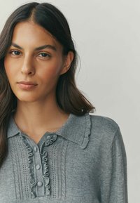 Gray knitted sweater with a collared neckline, ruffle accents, and button details; features vertical stitch patterns and soft texture.