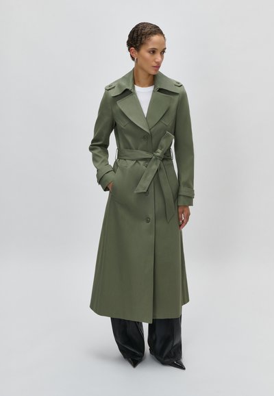 Γυναίκα που φοράει μακρύ χακί καμπαρντίνα με ζώνη πάνω από μαύρο παντελόνι με φαρδύ πόδι, στέκεται με το ένα χέρι στην τσέπη μπροστά σε απλό φόντο.