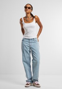 Hvit stripete tanktop, lyseblå baggy jeans, lagdelte sølvhalssmykker og brune joggesko med rosa lisser; modellen poserer mot en ensfarget bakgrunn.