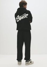 Black hoodie with a large white graphic on the back, paired with black pants and gray/white sneakers. Simple, relaxed-fit design.