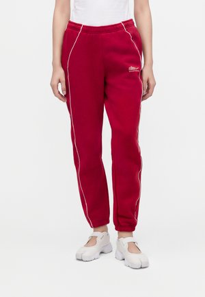 Personne portant un pantalon de survêtement rouge avec un liseré blanc et des baskets blanches, debout devant un fond clair uni.