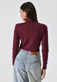 Bordeaux turtleneck trui met lange mouwen en knoopaccenten, gecombineerd met lichtblauwe high-waisted denim jeans met een leren label.