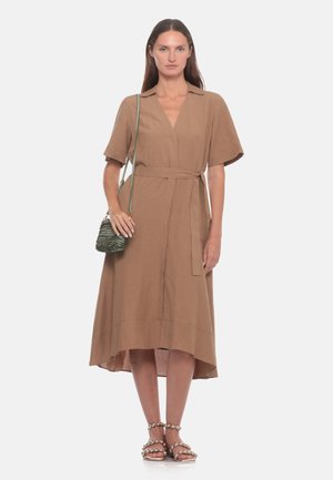 Donna che indossa un abito avvolgente marrone al ginocchio con maniche corte, porta una piccola borsa verde a tracolla e indossa sandali beige con perline.