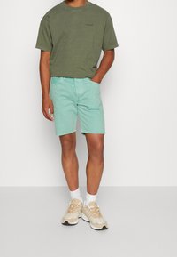 Camiseta de algodón en verde oliva combinada con pantalones cortos de mezclilla en verde menta. Calcetines blancos hasta el tobillo y zapatillas beis completan el atuendo. Diseño simple y casual.