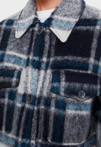 Kostkovaná flanelová košile v šedé, černé a modré barvě. Obsahuje límeček, dvě náprsní kapsy a knoflíky. Má texturovaný, jemný vzhled.