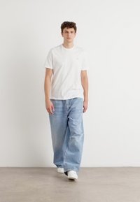 Camiseta blanca de algodón, jeans de mezclilla azul claro de corte holgado y zapatillas blancas. Modelo de pie contra un fondo claro y liso.