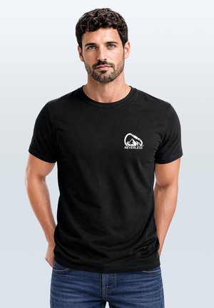 Mann mit dunklem lockigem Haar und Bart, der ein schwarzes T-Shirt mit weißem Berg- und Karabiner-Logo und dem Wort "NEVERLESS" auf der Brust trägt.