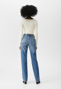 Top de manga larga de color beige claro con efecto acanalado, combinado con jeans de mezclilla azul de cintura alta. Los jeans tienen dos bolsillos traseros y un parche de cuero en la cinturilla.